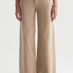 A person wearing beige wide-leg pants with a straight-to-slightly-flared leg, paired with a dark brown top and dark flat shoes, shown from the waist down against a light gray studio background.