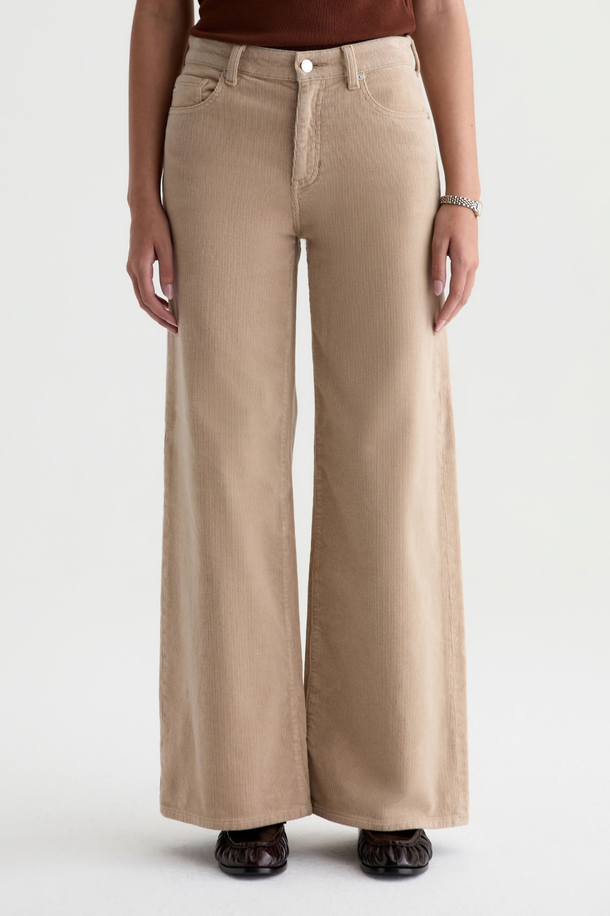 A person wearing beige wide-leg pants with a straight-to-slightly-flared leg, paired with a dark brown top and dark flat shoes, shown from the waist down against a light gray studio background.