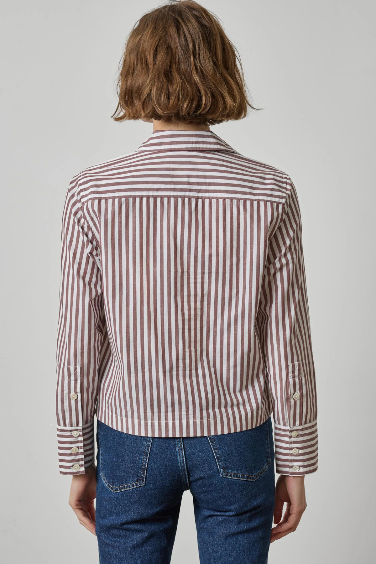 A person shown from the back wearing a cropped long-sleeve button-up shirt with vertical brown and white stripes, paired with blue jeans, against a light gray studio background.