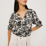 Woman wearing a black and white floral blouse with white pants on a plain background