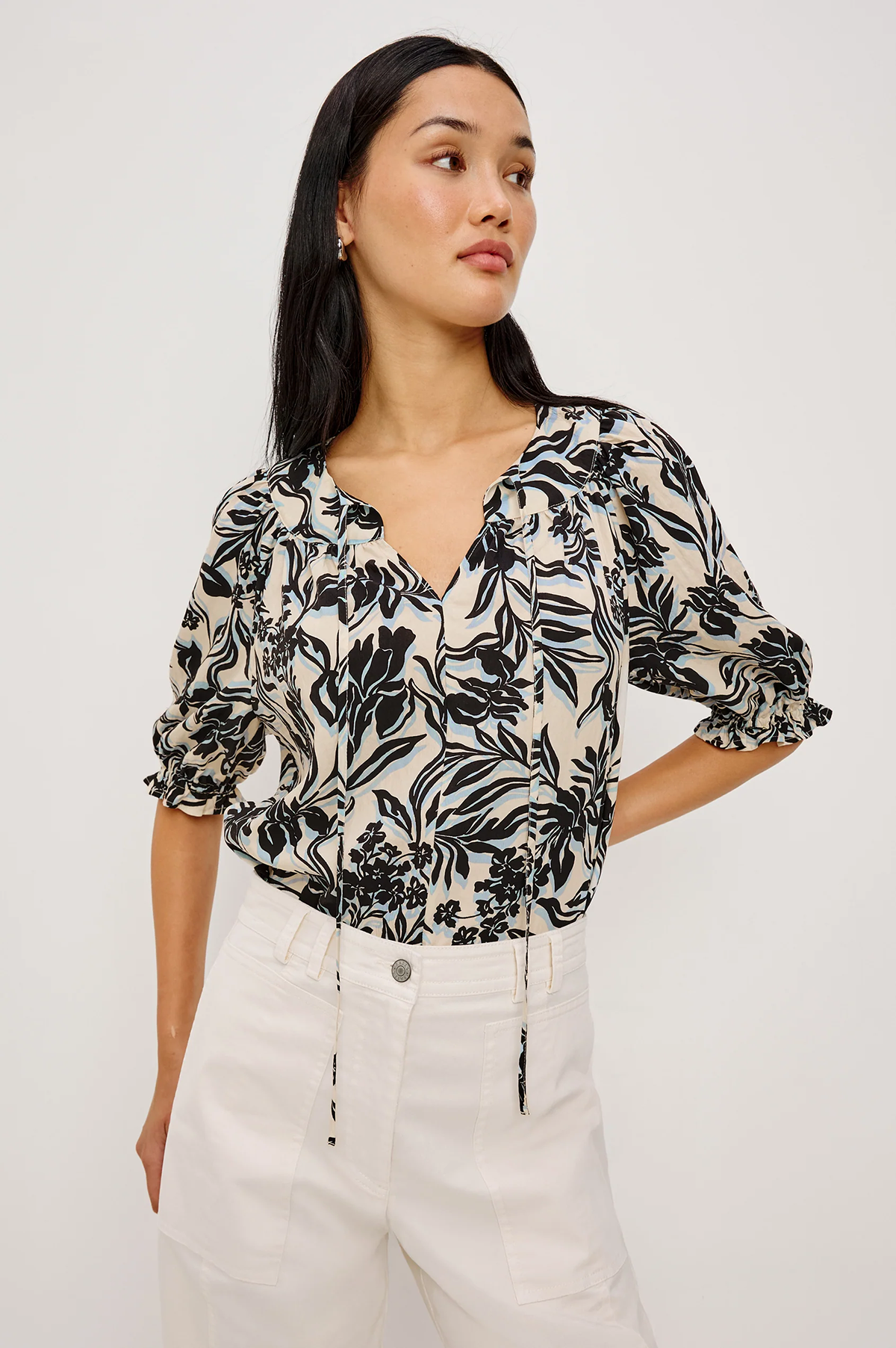 Woman wearing a black and white floral blouse with white pants on a plain background