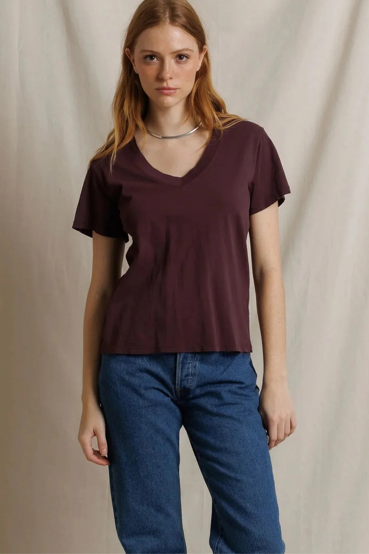 A woman modeling a short-sleeve V-neck top in a deep plum or burgundy shade, paired with blue jeans, standing against a neutral draped fabric backdrop.