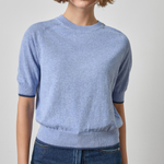 A woman wearing a light blue short-sleeve knit sweater with ribbed cuffs and hem, paired with dark blue jeans, photographed against a plain light gray studio background.