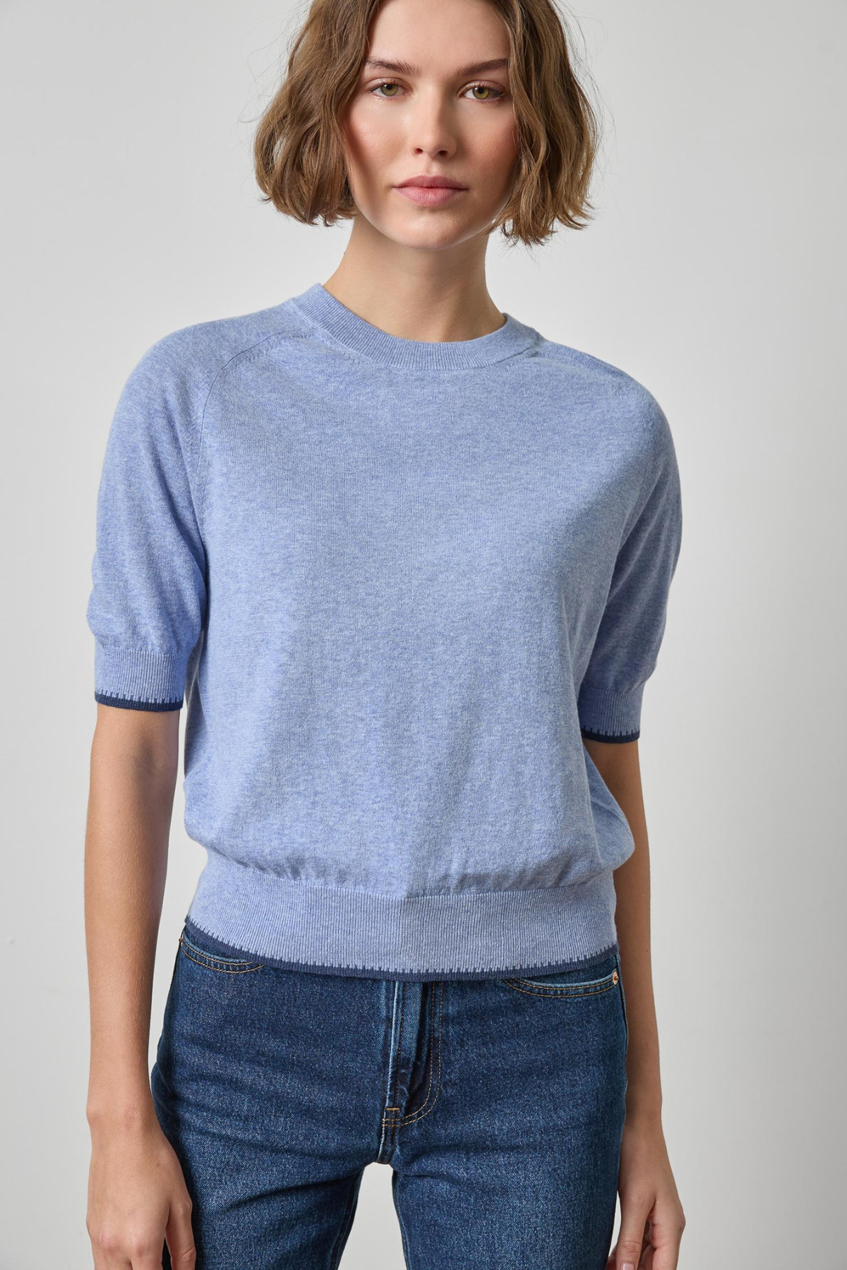 A woman wearing a light blue short-sleeve knit sweater with ribbed cuffs and hem, paired with dark blue jeans, photographed against a plain light gray studio background.
