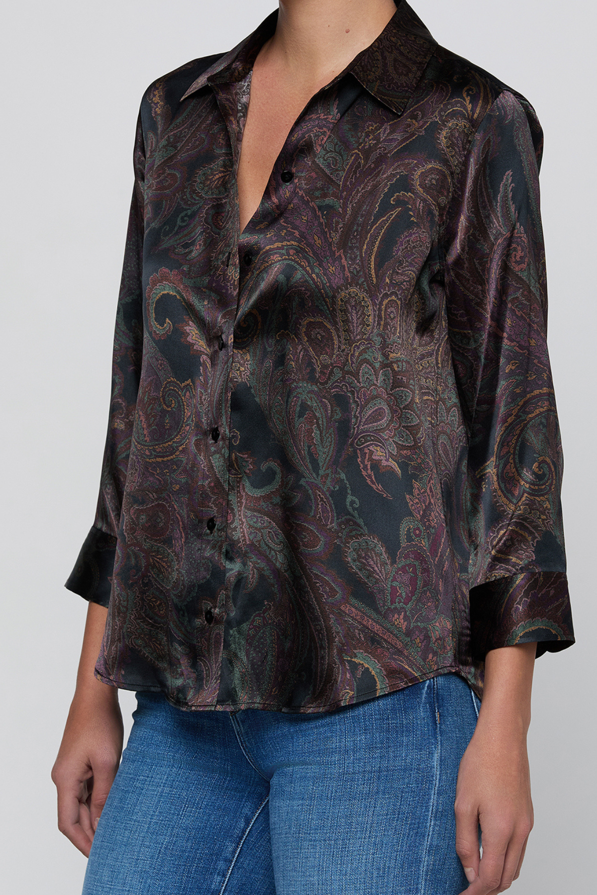 A woman wearing a dark paisley patterned button-up shirt with three-quarter sleeves and blue jeans, shown from the neck down against a light gray background.
