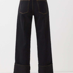 Rear view of a model wearing dark indigo wide-leg jeans with rolled cuffs and contrast gold-toned stitching, paired with a black top and brown boots against a white studio background.
