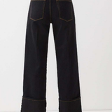 Rear view of a model wearing dark indigo wide-leg jeans with rolled cuffs and contrast gold-toned stitching, paired with a black top and brown boots against a white studio background.