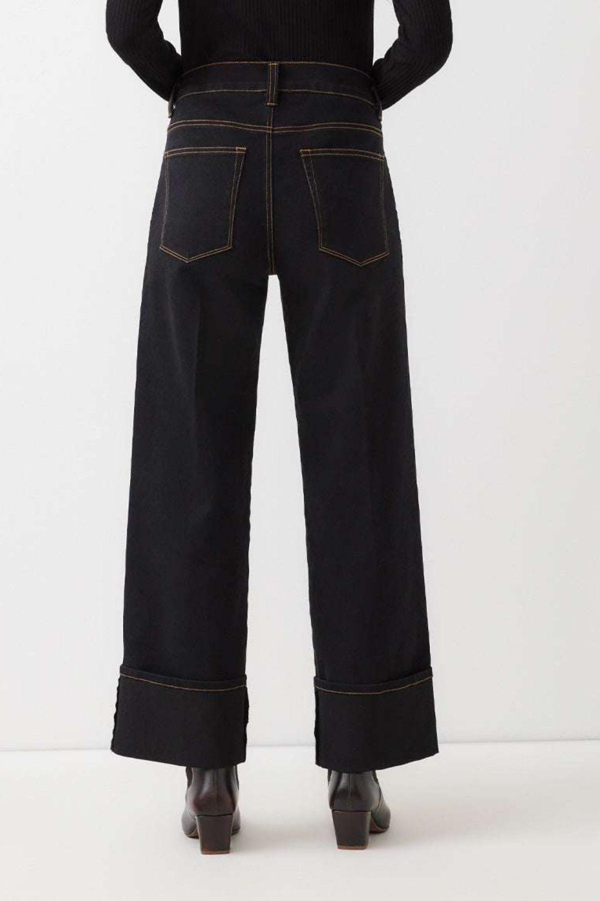 Rear view of a model wearing dark indigo wide-leg jeans with rolled cuffs and contrast gold-toned stitching, paired with a black top and brown boots against a white studio background.