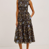 A woman modeling a sleeveless midi dress with a dark brown floral print, ruffled shoulders, and a gathered waist, paired with black strappy heels against a plain white studio background.