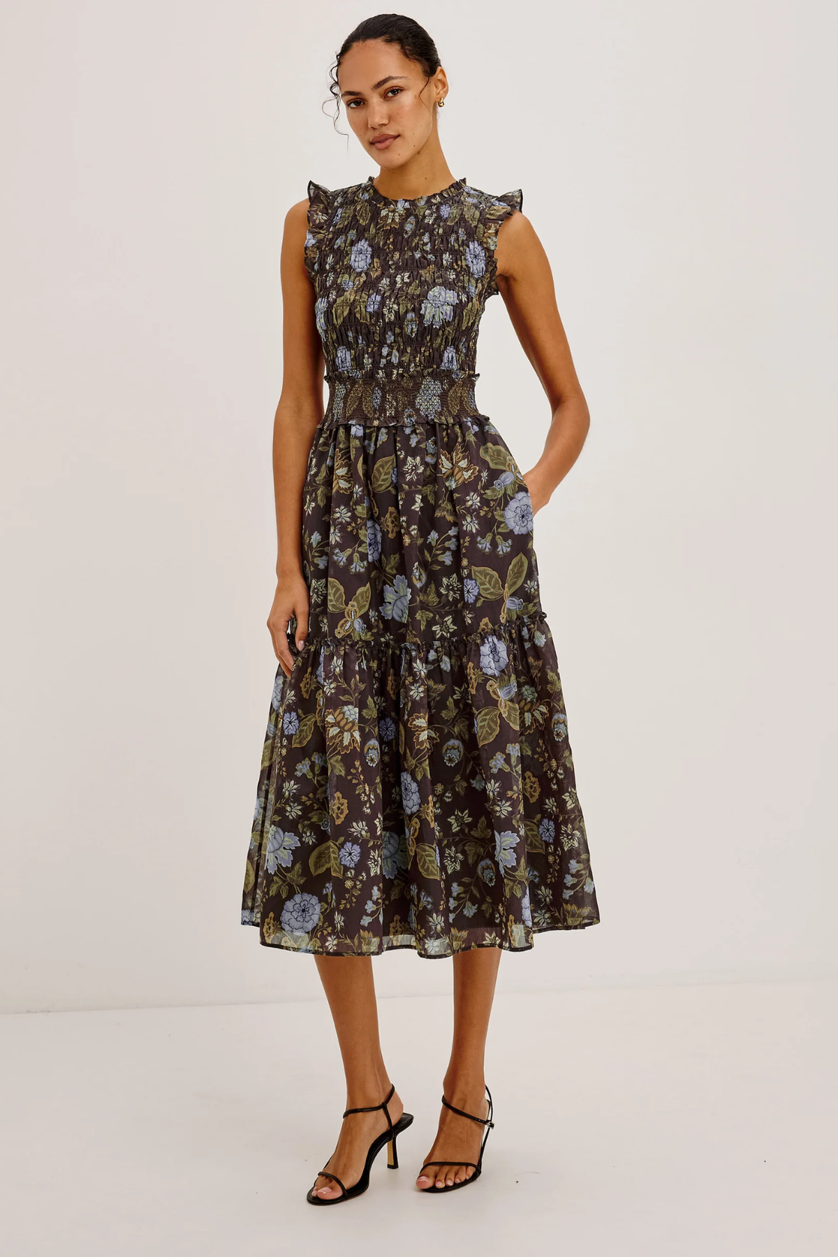 A woman modeling a sleeveless midi dress with a dark brown floral print, ruffled shoulders, and a gathered waist, paired with black strappy heels against a plain white studio background.