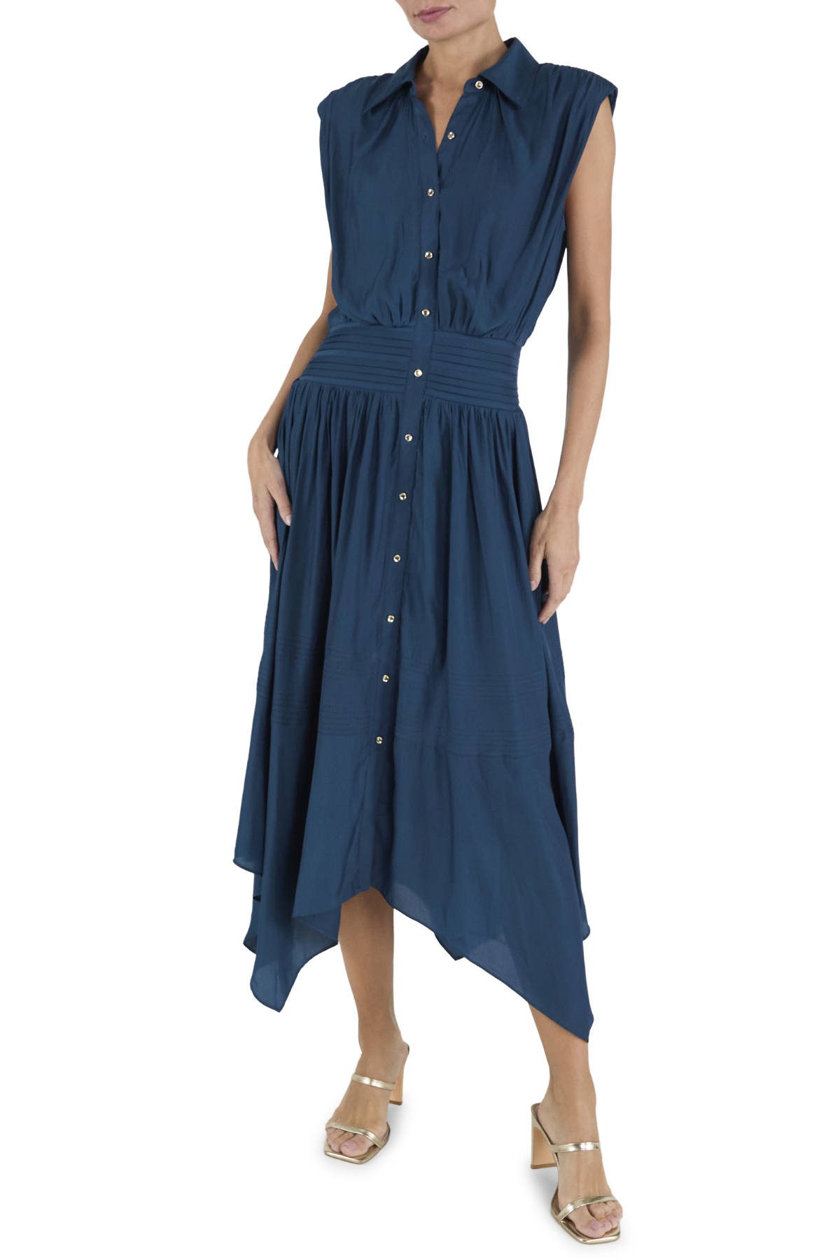 A woman modeling a sleeveless navy blue midi dress with a button-down front, smocked waist, and asymmetrical hem, paired with beige heeled sandals against a white background.