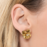 A close-up of a model's ear wearing a textured gold flower-shaped stud earring with a small clear center stone.