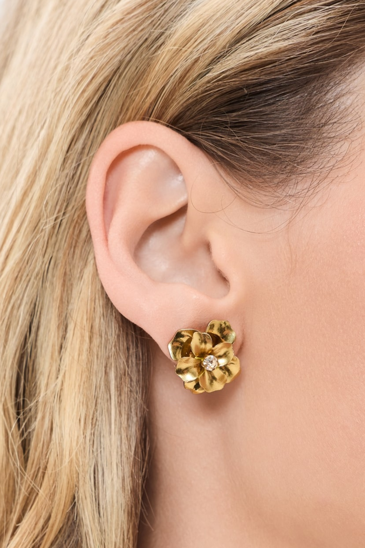 A close-up of a model's ear wearing a textured gold flower-shaped stud earring with a small clear center stone.