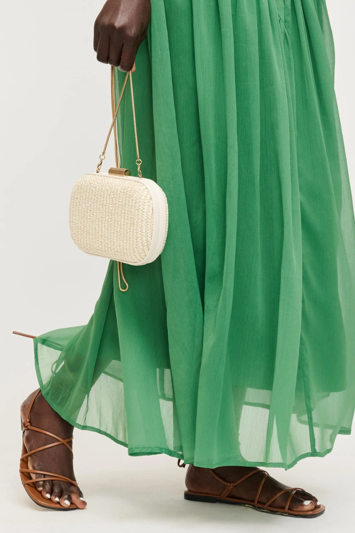 Person wearing a green dress and brown sandals holding a white woven handbag.
