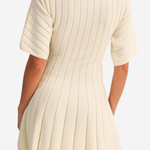 A woman wearing a cream-colored ribbed knit dress with pleated details, shown from the back against a plain white background.