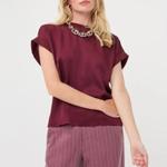 A woman modeling a short-sleeve burgundy top with a wide collar and matching mauve pinstriped trousers, accessorized with a chunky silver necklace, against a plain white studio background.