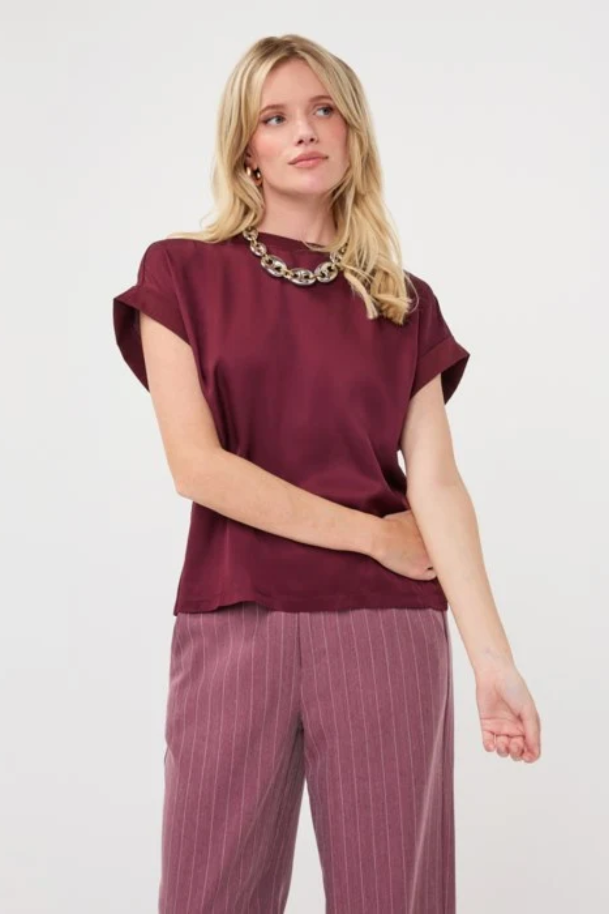 A woman modeling a short-sleeve burgundy top with a wide collar and matching mauve pinstriped trousers, accessorized with a chunky silver necklace, against a plain white studio background.