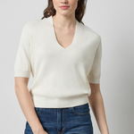 A woman modeling a short-sleeve cream knit V-neck sweater with blue jeans, photographed against a plain light gray studio background.