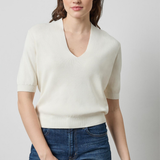 A woman modeling a short-sleeve cream knit V-neck sweater with blue jeans, photographed against a plain light gray studio background.