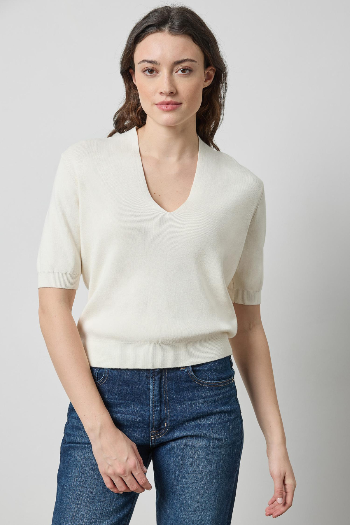 A woman modeling a short-sleeve cream knit V-neck sweater with blue jeans, photographed against a plain light gray studio background.