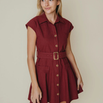 A woman modeling a short burgundy button-down shirt dress with a matching belt and gold buckle, set against a plain beige background.