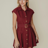 A woman modeling a short burgundy button-down shirt dress with a matching belt and gold buckle, set against a plain beige background.