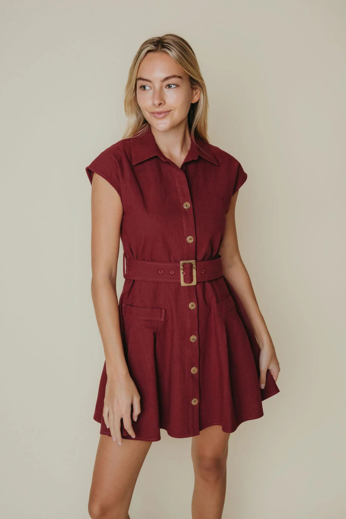A woman modeling a short burgundy button-down shirt dress with a matching belt and gold buckle, set against a plain beige background.