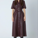 A woman modeling a dark burgundy faux-leather shirt dress with short sleeves, a button-down front, and a matching belt with a silver buckle, paired with black knee-high boots against a light gray studio background.