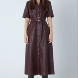 A woman modeling a dark burgundy faux-leather shirt dress with short sleeves, a button-down front, and a matching belt with a silver buckle, paired with black knee-high boots against a light gray studio background.