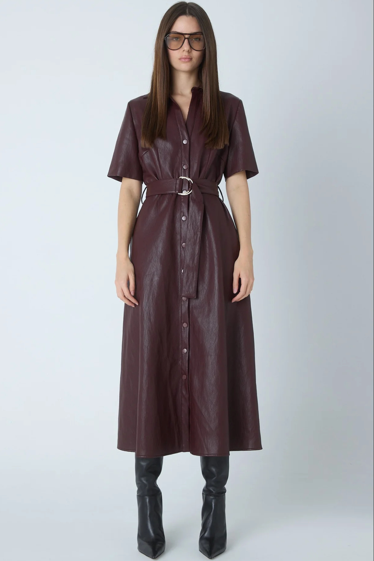 A woman modeling a dark burgundy faux-leather shirt dress with short sleeves, a button-down front, and a matching belt with a silver buckle, paired with black knee-high boots against a light gray studio background.