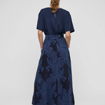 A woman viewed from behind wearing a navy blue short-sleeve maxi dress with a textured floral jacquard skirt and black strappy heels, set against a plain light gray studio background.