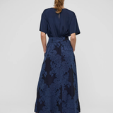 A woman viewed from behind wearing a navy blue short-sleeve maxi dress with a textured floral jacquard skirt and black strappy heels, set against a plain light gray studio background.