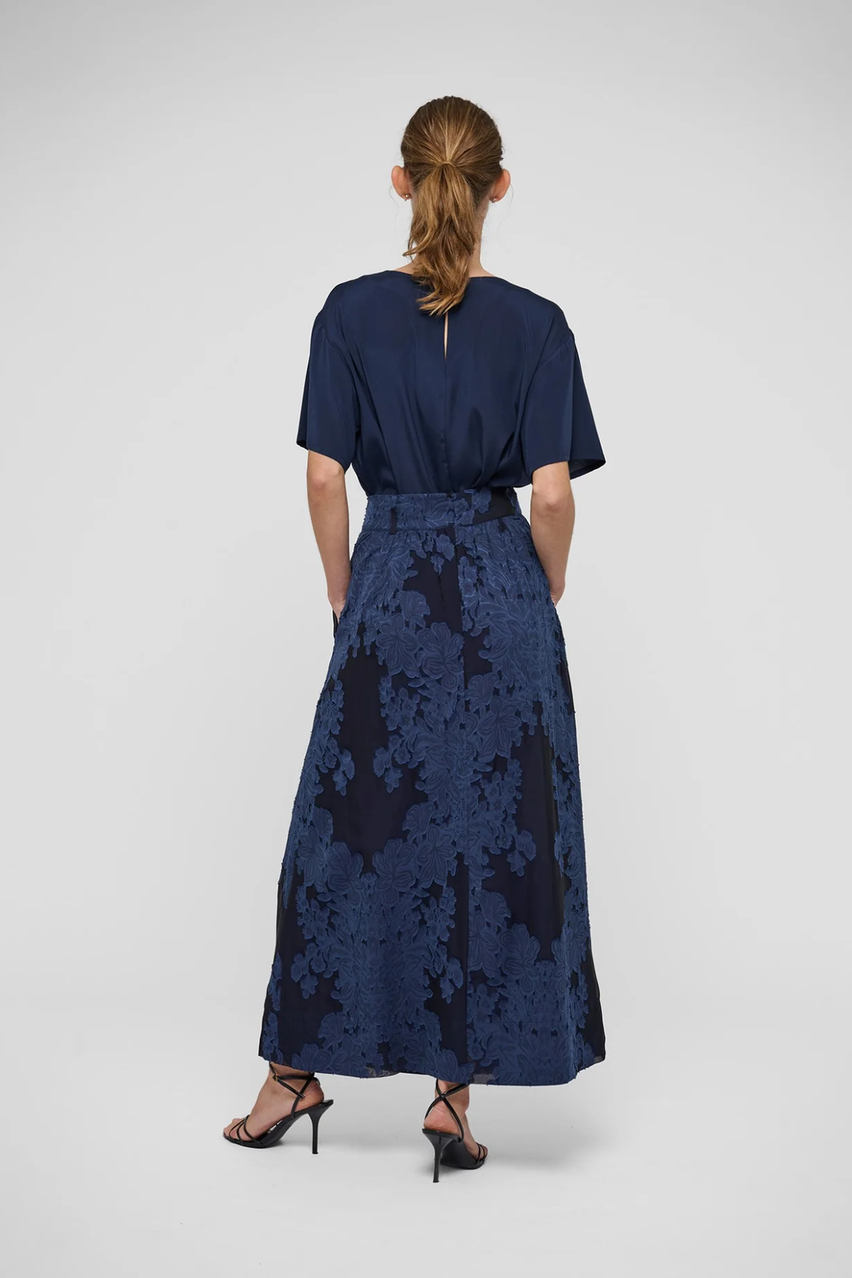 A woman viewed from behind wearing a navy blue short-sleeve maxi dress with a textured floral jacquard skirt and black strappy heels, set against a plain light gray studio background.