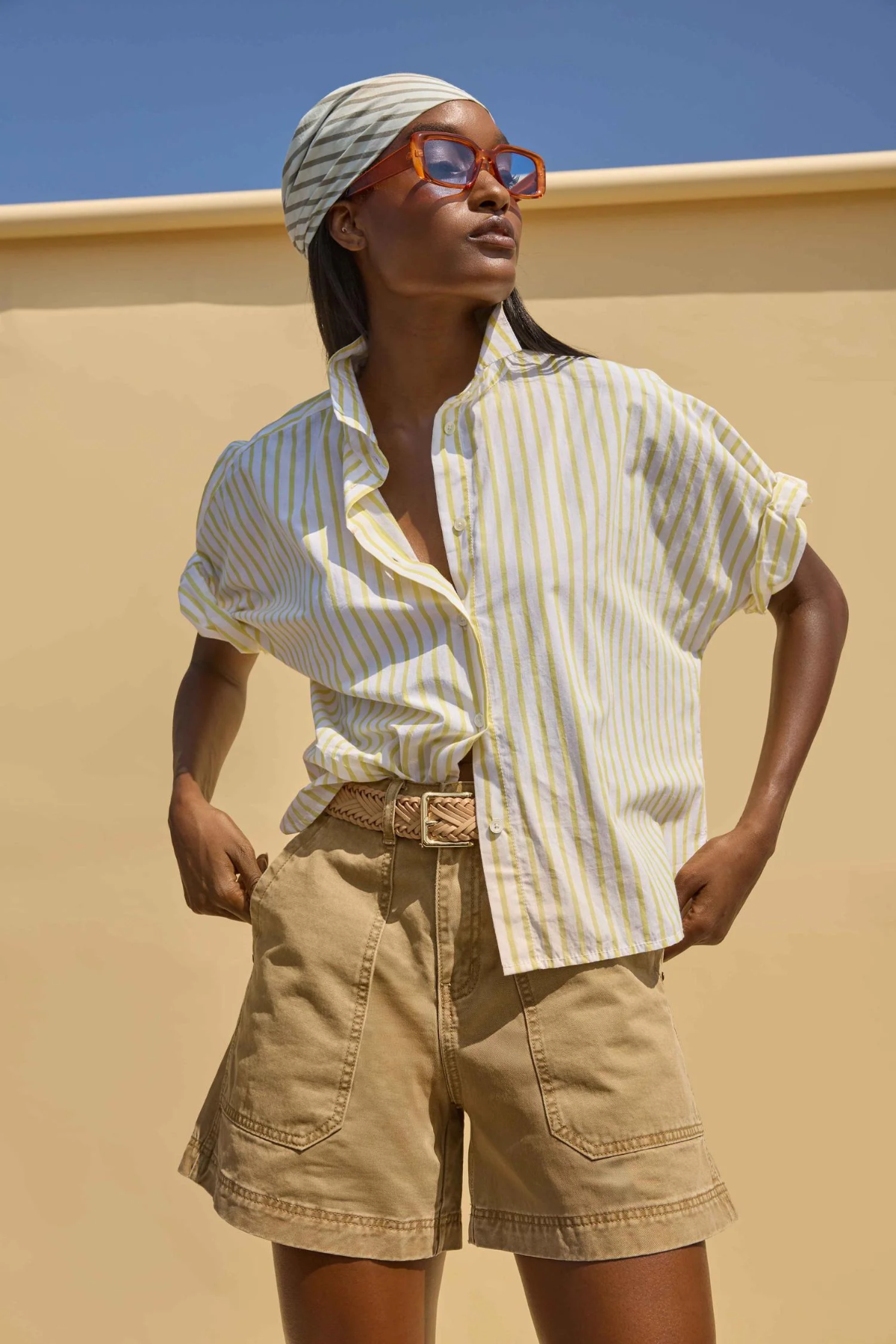 Woman wearing a striped shirt and shorts against a beige wall with blue sky.