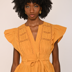A woman with a large black afro wearing a mustard yellow peplum blouse with ruffled cap sleeves, a V-neckline, and decorative chain-like embroidery on the chest, paired with dark brown velvet pants against a plain light beige background.