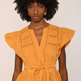 A woman with a large black afro wearing a mustard yellow peplum blouse with ruffled cap sleeves, a V-neckline, and decorative chain-like embroidery on the chest, paired with dark brown velvet pants against a plain light beige background.