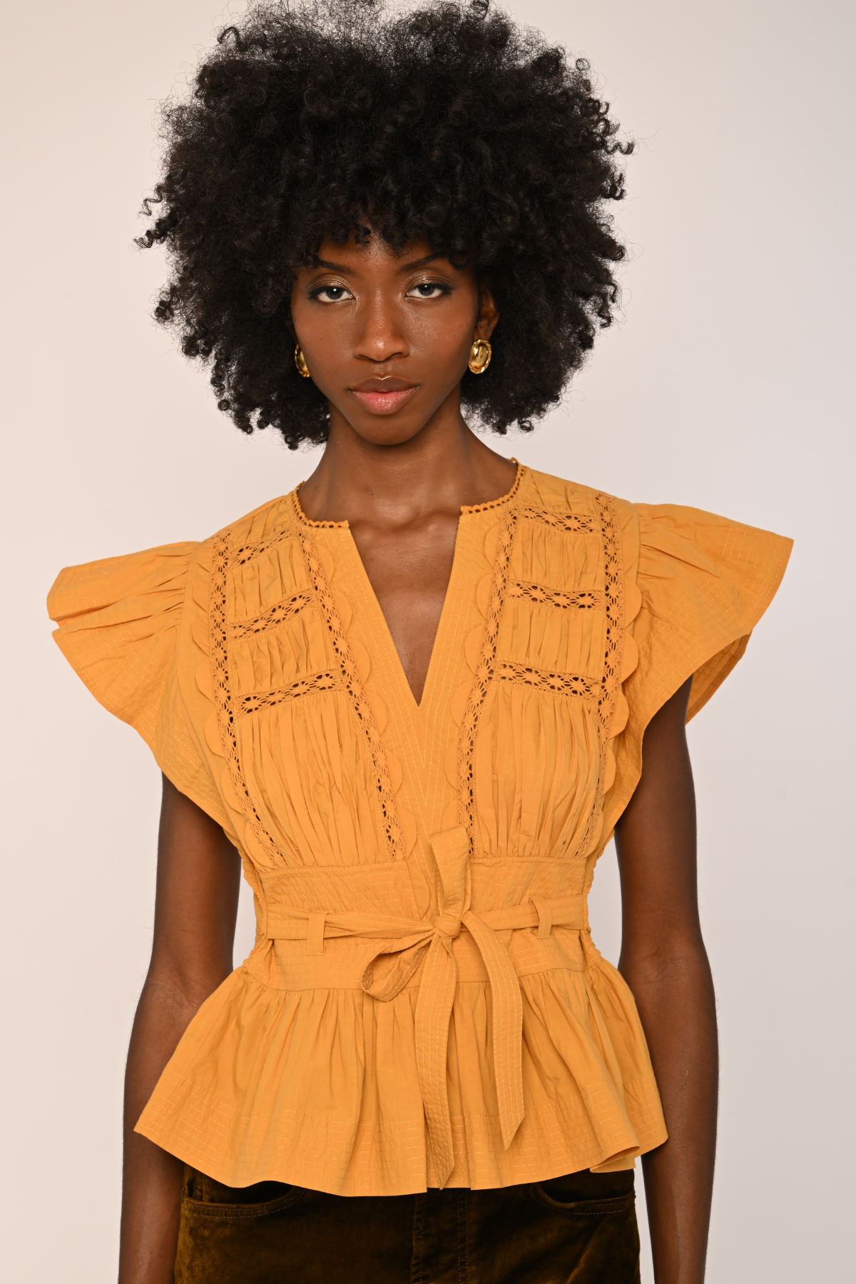 A woman with a large black afro wearing a mustard yellow peplum blouse with ruffled cap sleeves, a V-neckline, and decorative chain-like embroidery on the chest, paired with dark brown velvet pants against a plain light beige background.