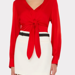 A woman wearing a bright red long-sleeve wrap-style top with a front knot detail and a white mini skirt with a thin black belt, photographed against a light gray studio background.