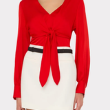 A woman wearing a bright red long-sleeve wrap-style top with a front knot detail and a white mini skirt with a thin black belt, photographed against a light gray studio background.