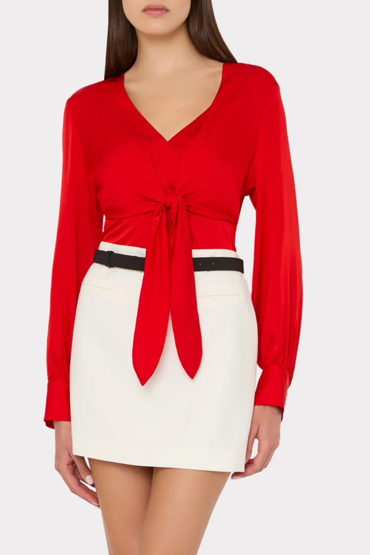 A woman wearing a bright red long-sleeve wrap-style top with a front knot detail and a white mini skirt with a thin black belt, photographed against a light gray studio background.