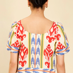 A woman viewed from the back wearing a short-sleeve dress with a cream base and a bold red, blue, and yellow ikat pattern, set against a plain beige background.