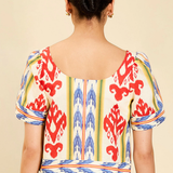 A woman viewed from the back wearing a short-sleeve dress with a cream base and a bold red, blue, and yellow ikat pattern, set against a plain beige background.
