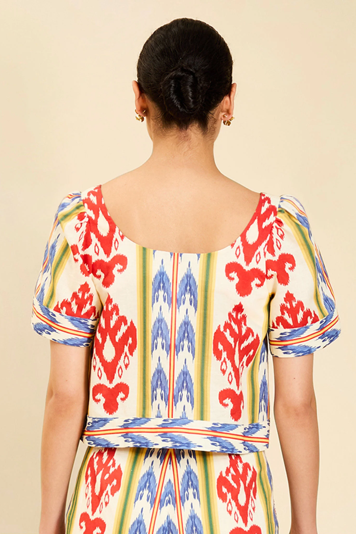 A woman viewed from the back wearing a short-sleeve dress with a cream base and a bold red, blue, and yellow ikat pattern, set against a plain beige background.