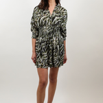A woman modeling a short wrap-style dress with three-quarter sleeves, featuring an abstract black, white, and olive green pattern, paired with beige slide sandals against a light gray studio background.