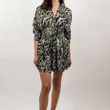A woman modeling a short wrap-style dress with three-quarter sleeves, featuring an abstract black, white, and olive green pattern, paired with beige slide sandals against a light gray studio background.