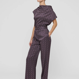 A woman modeling a dark purple and black geometric patterned jumpsuit with a mock neck and wide-leg pants, standing against a light gray studio background.