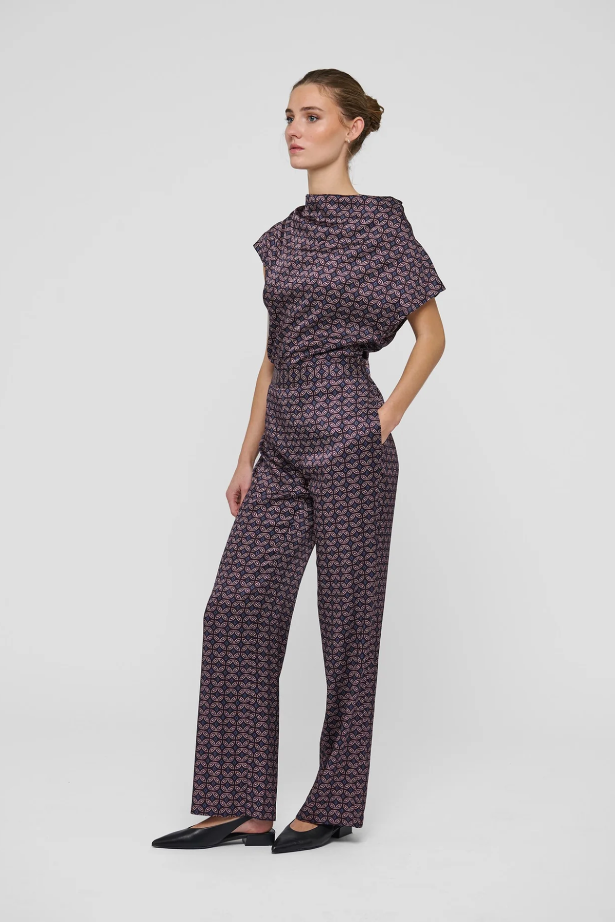 A woman modeling a dark purple and black geometric patterned jumpsuit with a mock neck and wide-leg pants, standing against a light gray studio background.