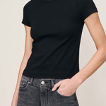 A woman wearing a black short-sleeve crewneck t-shirt and dark gray jeans, shown from the neck down against a plain light beige background.