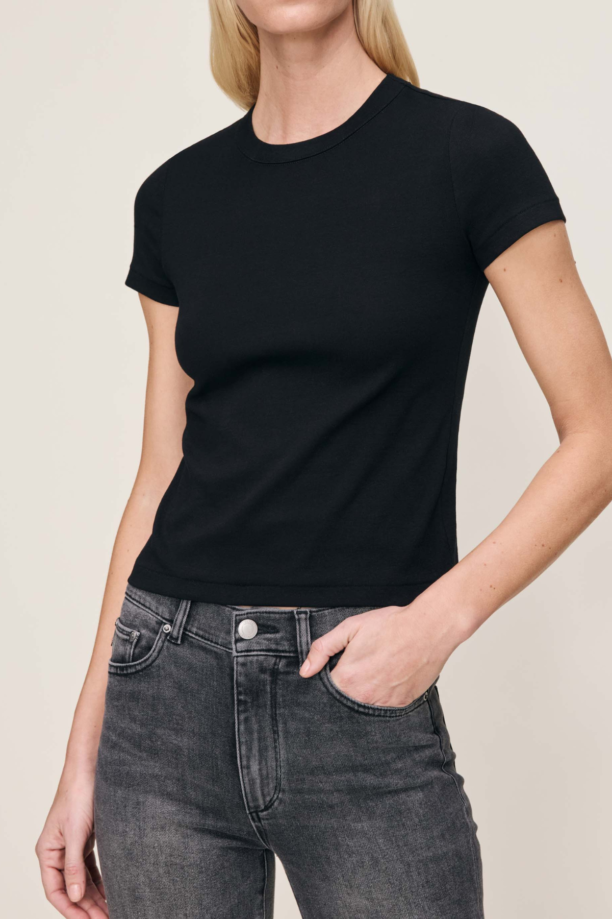 A woman wearing a black short-sleeve crewneck t-shirt and dark gray jeans, shown from the neck down against a plain light beige background.