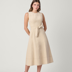 A woman modeling a sleeveless, knee-length beige dress with a matching waist tie, paired with brown ankle boots, against a light gray studio background.